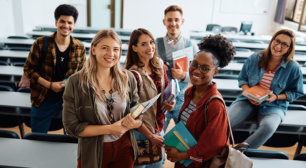 Group of six diverse and smiling university students standing together in a classroom, holding books and notebooks, with rows of desks and chairs in the background.